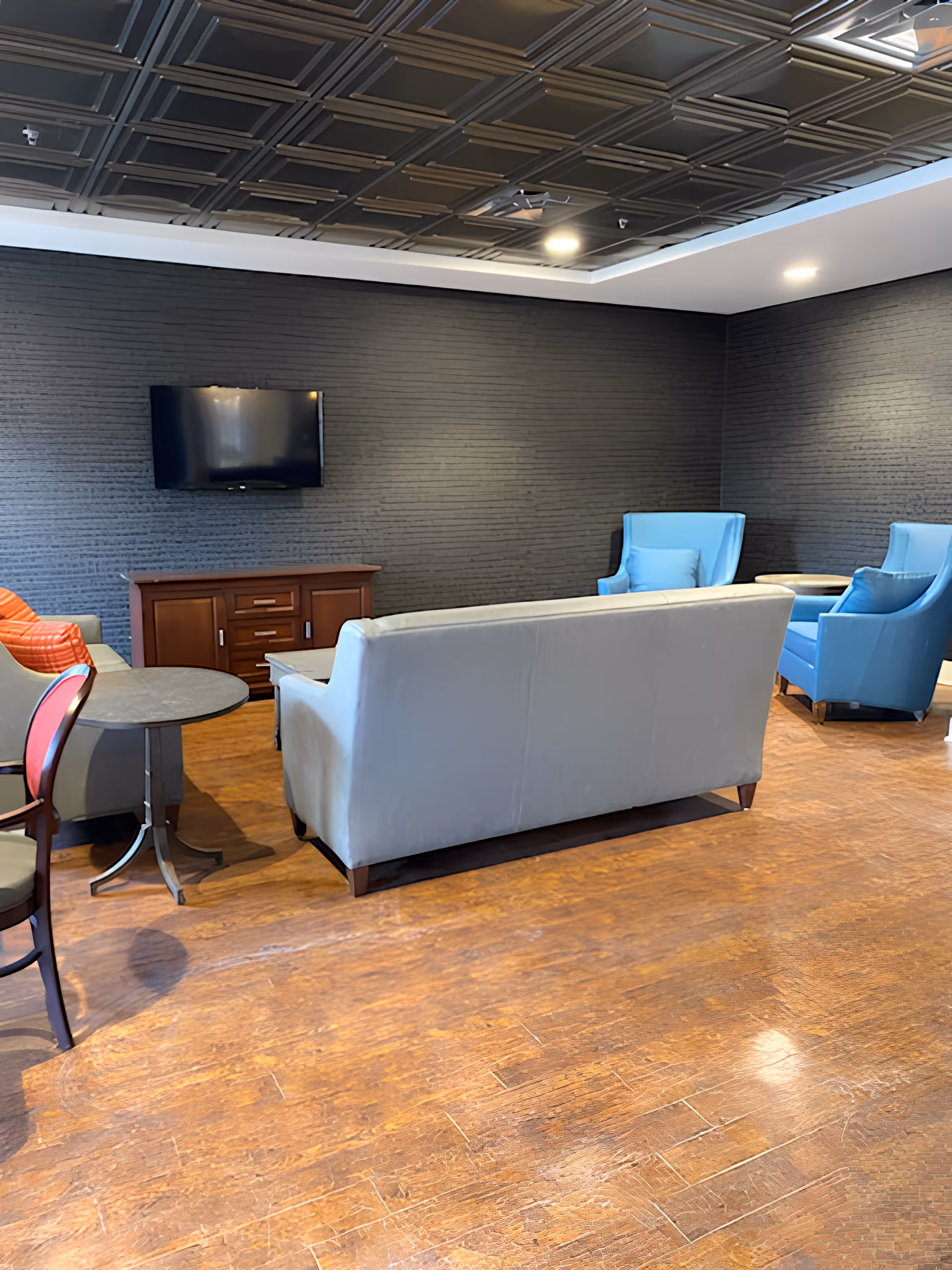 A senior living facility common area with a gray sofa facing a wall-mounted flat screen TV above a wooden cabinet. There are two blue armchairs with matching pillows and a small round table between them. The floor is wooden, and the walls are dark textured brick. The ceiling has a decorative pattern with recessed lighting.