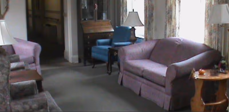 A communal living room with sofas, armchairs, lamps, and side tables near windows.