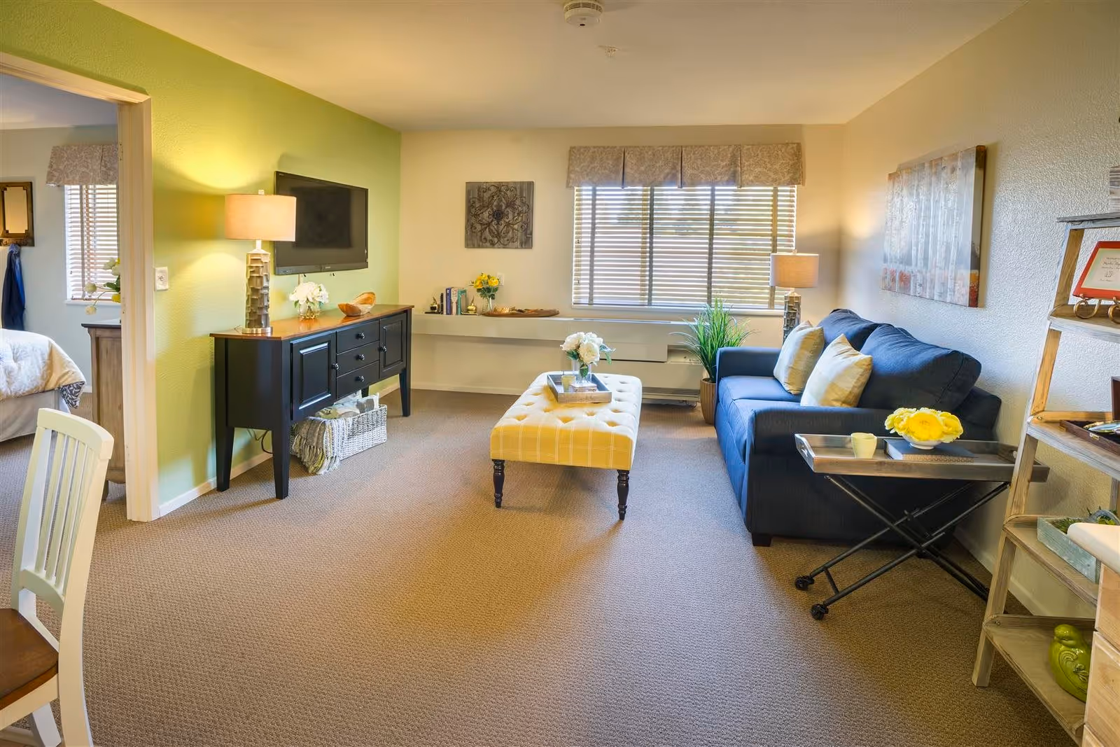 A bright and cozy living room with a blue sofa adorned with yellow and white pillows, a yellow tufted ottoman in the center, and a black console table with a lamp and decorative items against a green accent wall. A window with blinds lets in natural light, and a small wooden shelf holds framed pictures and decor. A doorway leads to a bedroom with a bed partially visible.
