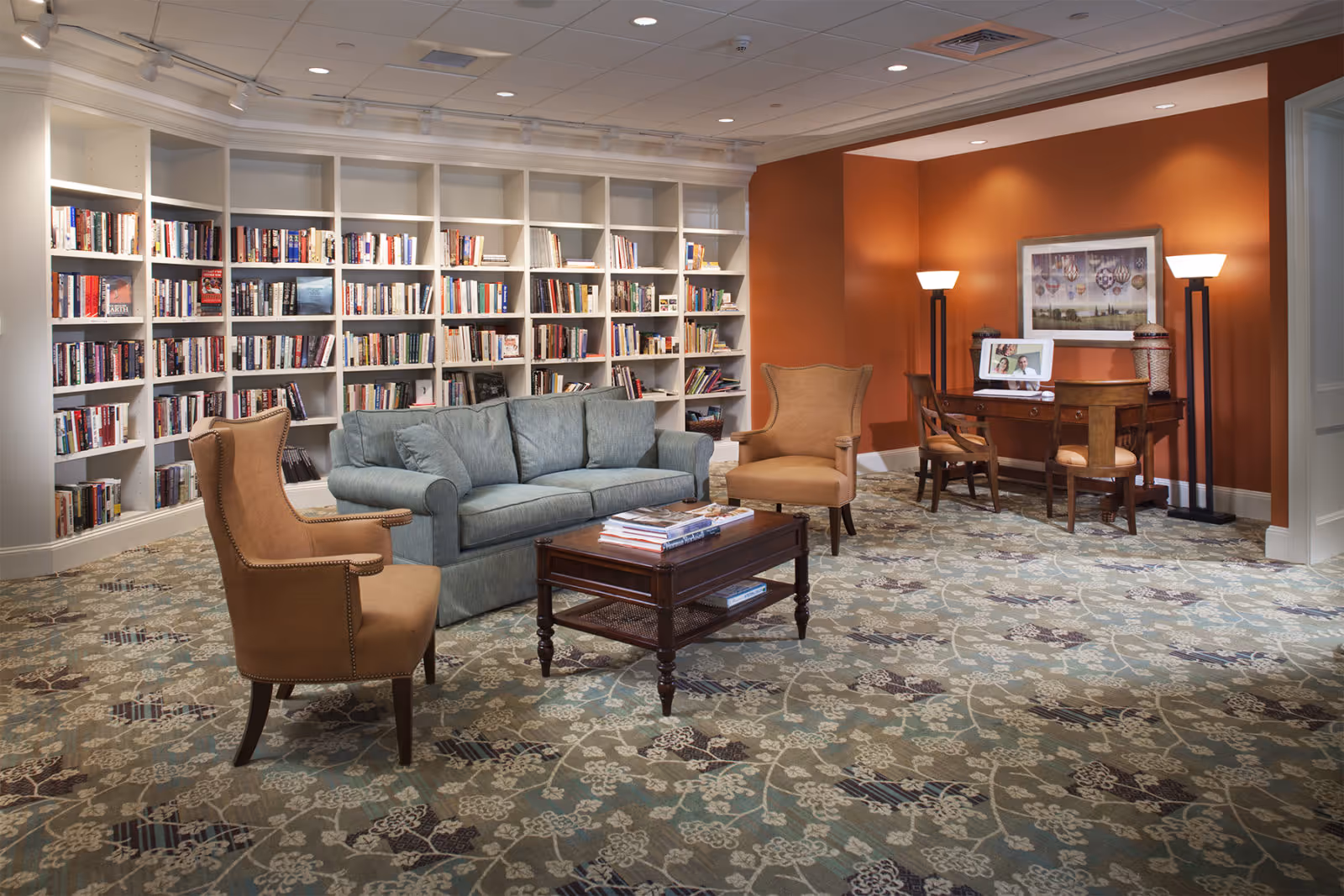 A cozy library or lounge area with a large white bookshelf filled with books along the back wall. In front of the bookshelf is a blue sofa and two tan armchairs around a wooden coffee table with books on it. To the right, there is a wooden desk with two chairs, a digital photo frame, and two tall floor lamps against an orange accent wall with a framed picture above the desk. The floor is covered with a patterned carpet.