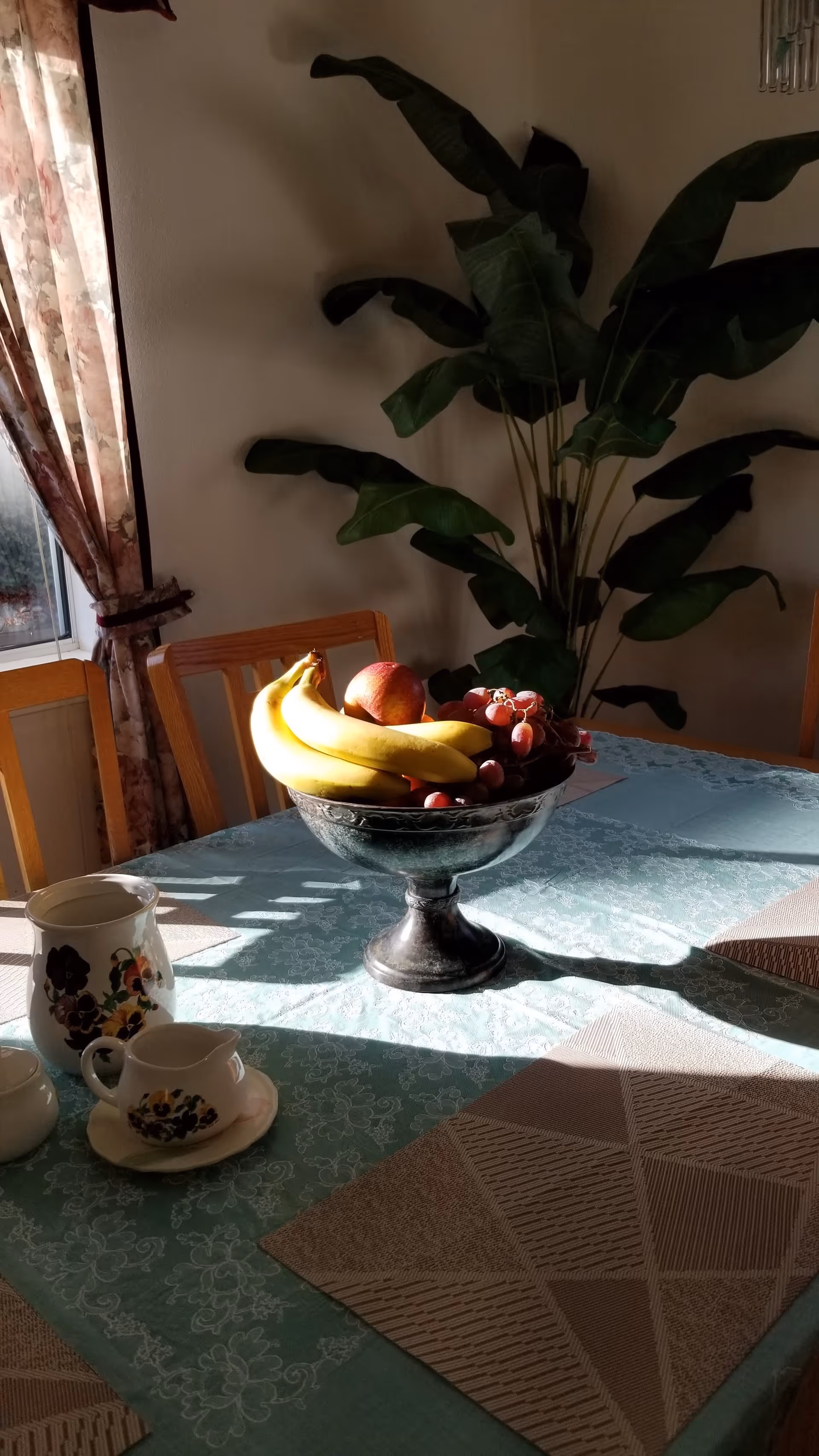 A dining table with a green floral tablecloth and beige placemats, featuring a silver fruit bowl filled with bananas, grapes, and an apple. There are also a floral-patterned pitcher, creamer, and sugar bowl on the table. In the background, there is a large green leafy plant and a window with floral curtains letting in sunlight.