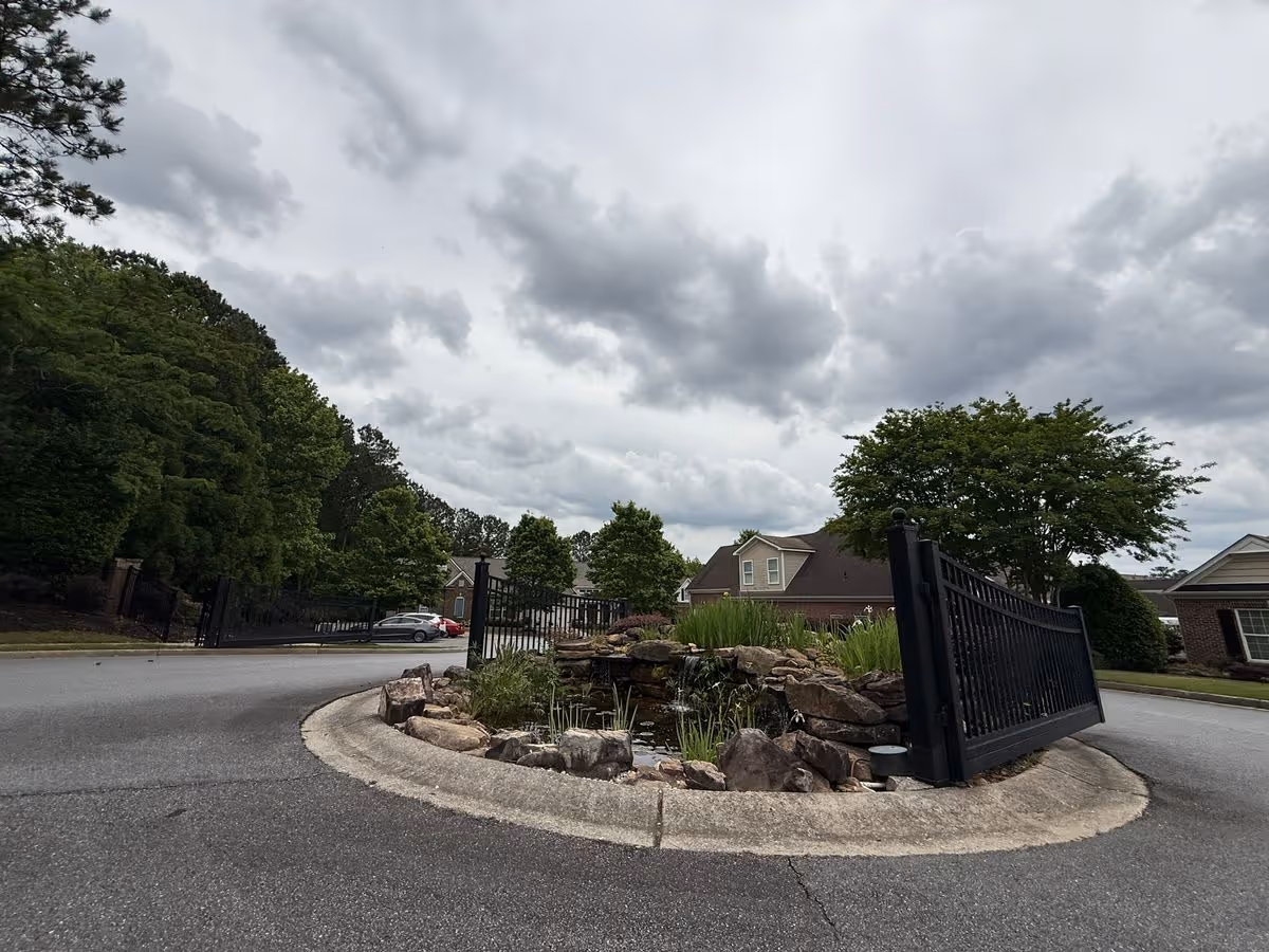 Gated community entrance with a stone-lined pond and decorative black gate in front of houses under a cloudy sky.