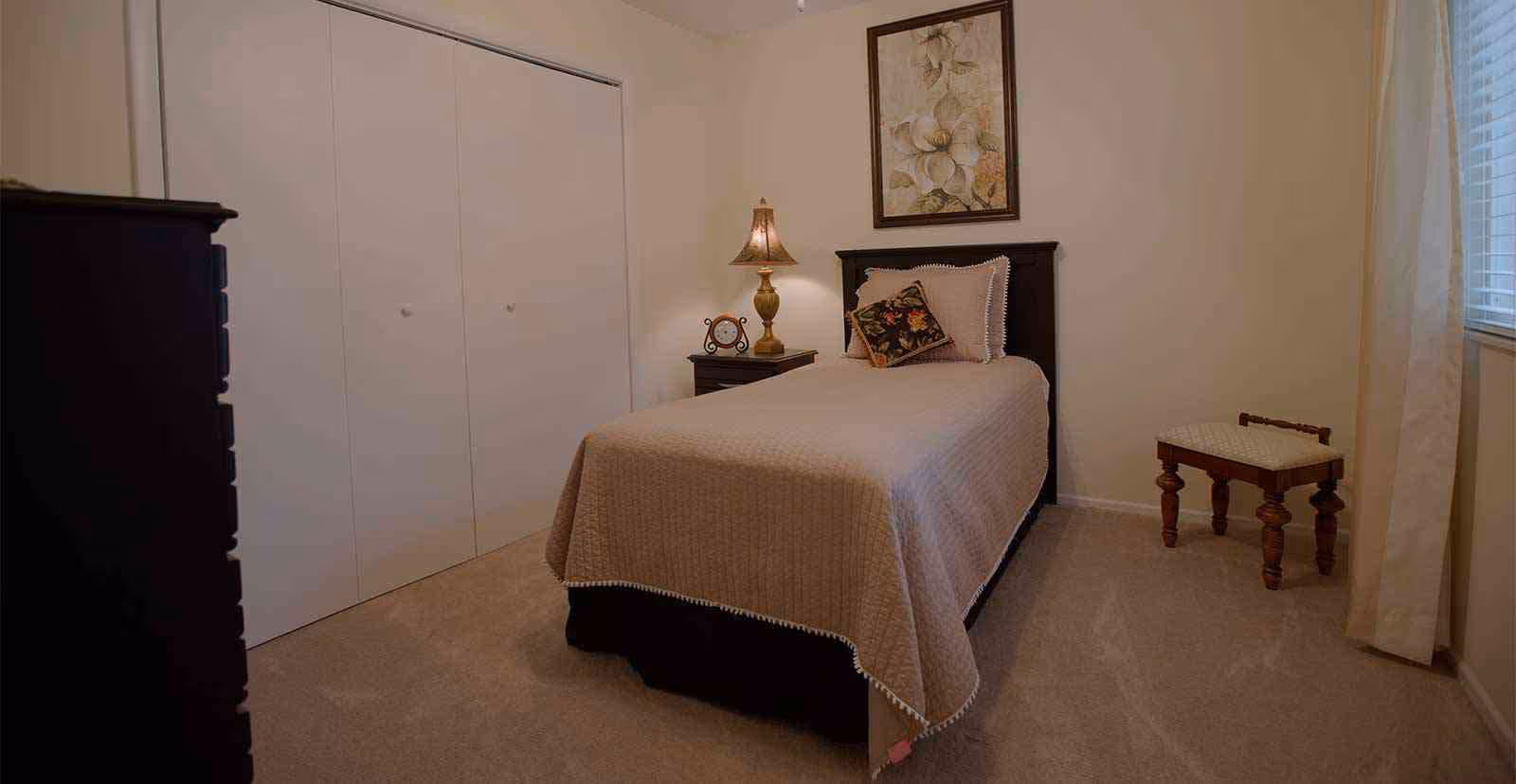 A small bedroom with a single bed covered in a beige quilt and a decorative pillow. There is a wooden nightstand with a lamp and a clock next to the bed. A framed floral artwork hangs above the bed. A small wooden stool is placed near a window with white curtains. The room has beige carpet and white walls with a closed closet door.