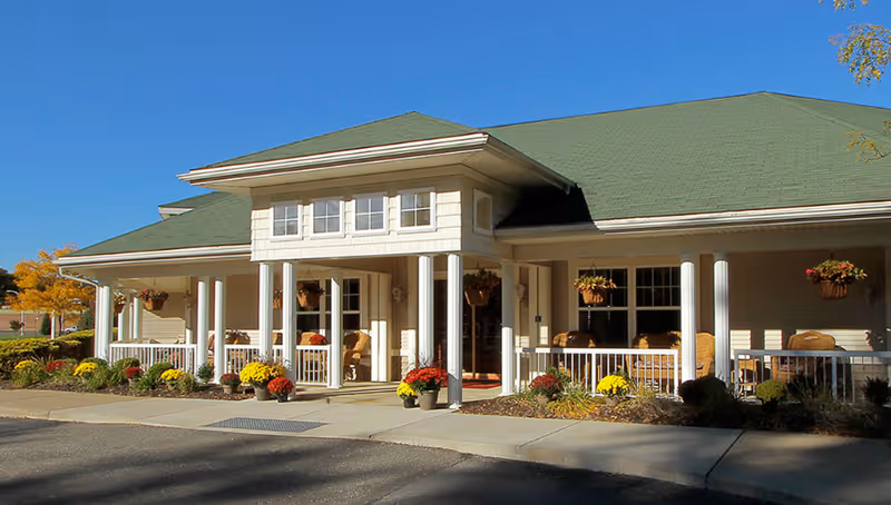 Front exterior of a single-story senior living facility with a covered porch, white columns, potted flowers, and a green roof under a clear blue sky.