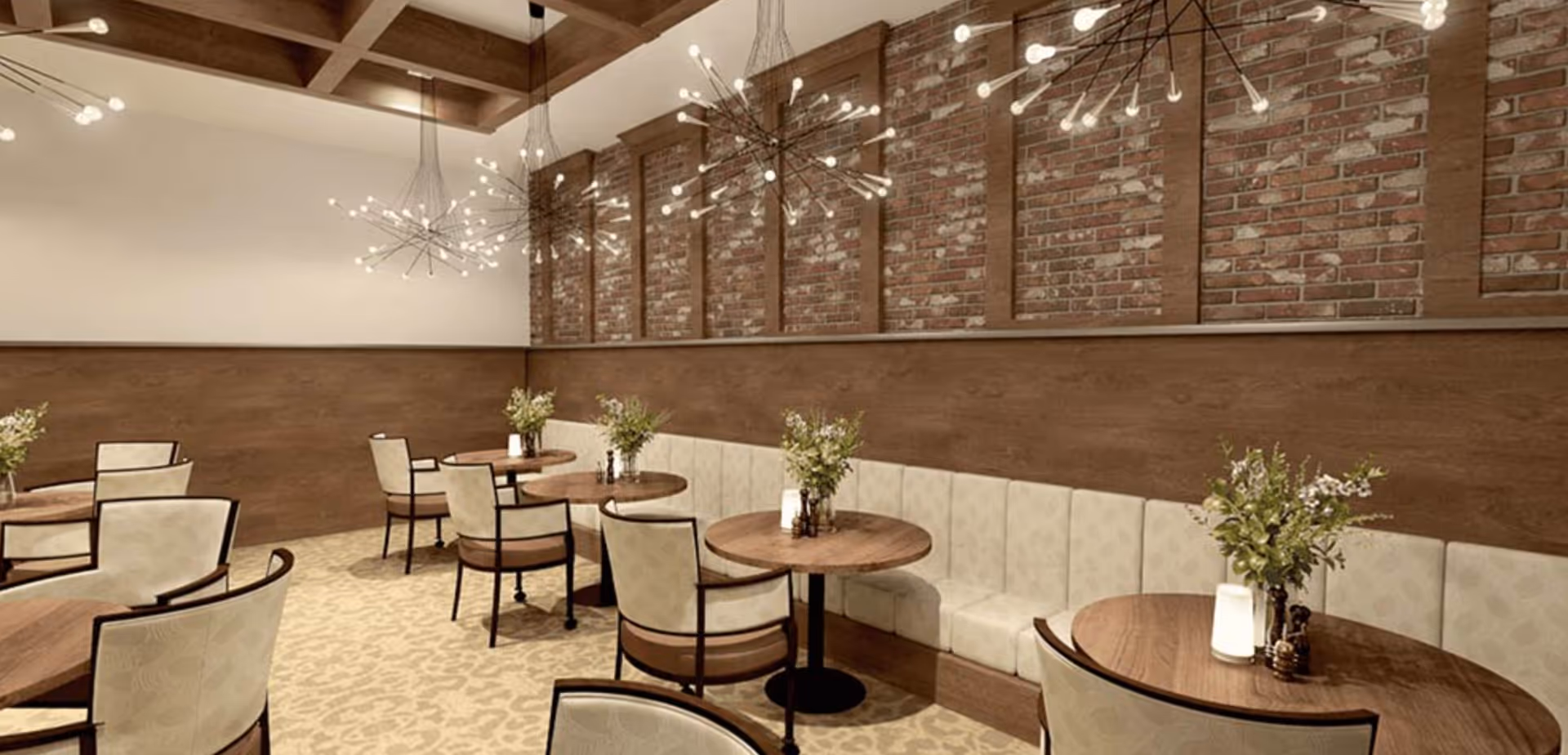 A dining area with round wooden tables and cushioned chairs arranged neatly. The room features a long upholstered bench along a wall with exposed brick and wood paneling. Modern chandelier lights hang from a coffered ceiling, and small floral arrangements and candles decorate each table.