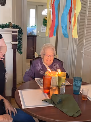 An elderly woman sitting at a dining table with lit birthday candles on a cake in front of her. Colorful streamers hang from the ceiling above her. There are drinking glasses and a salt shaker on the table. The setting appears to be a cozy indoor dining area with a door and some decorations in the background.