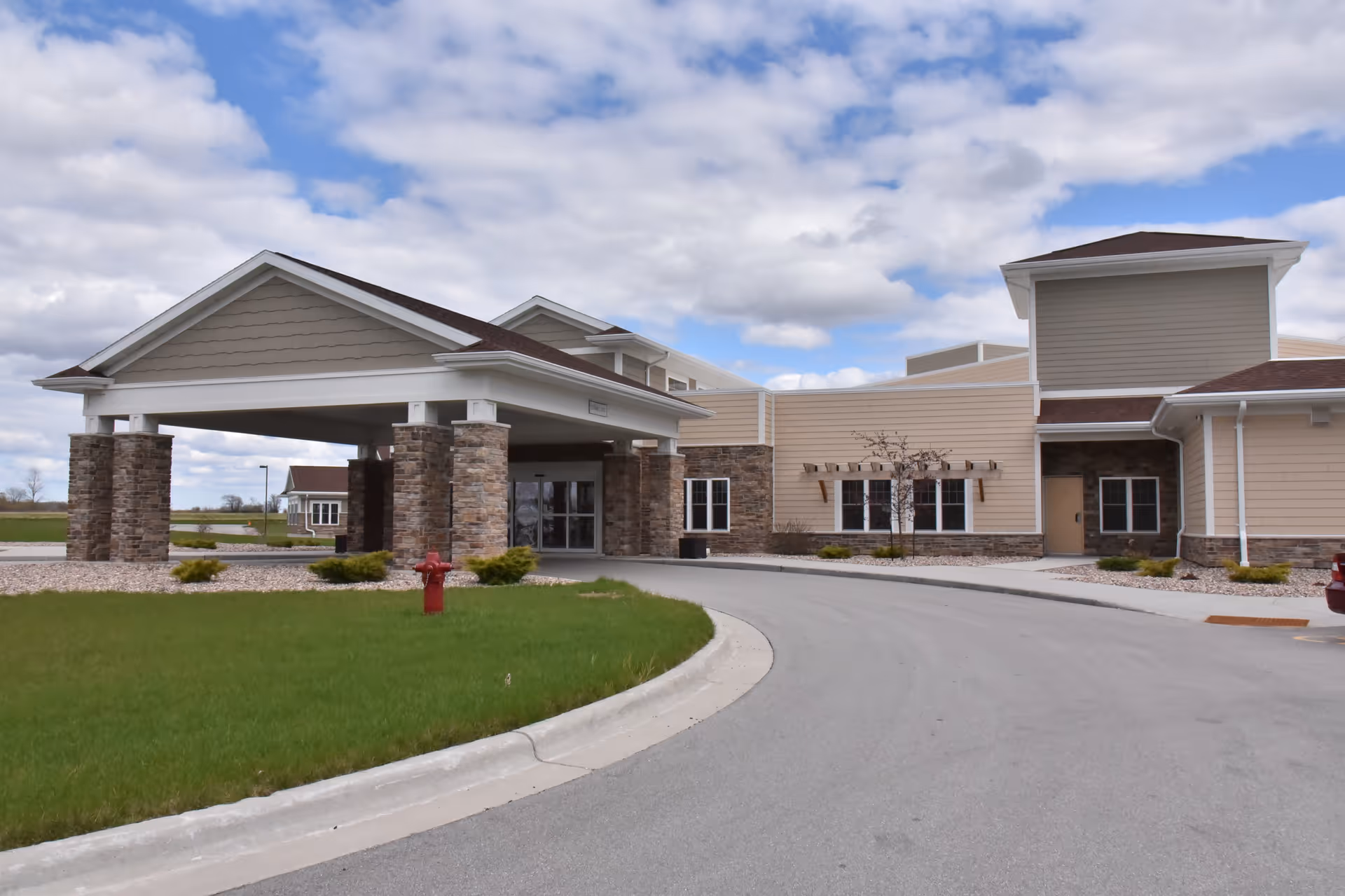 Front entrance and porte-cochère of a beige senior living building with stone columns and a curved driveway under a partly cloudy sky.