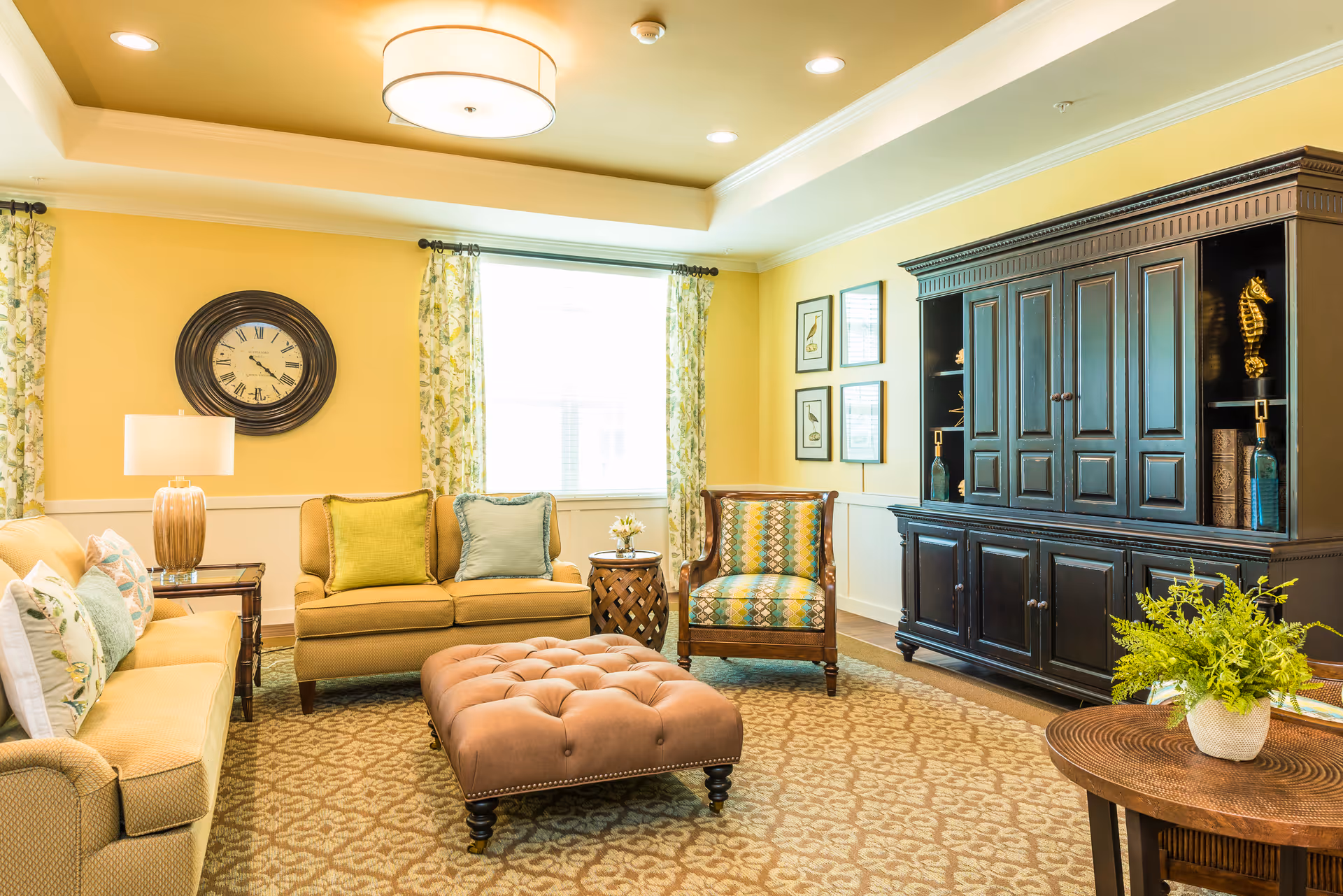 Bright, spacious living room with yellow walls featuring sofas, an upholstered ottoman, an armchair, a large dark wood cabinet, and decorative accents.