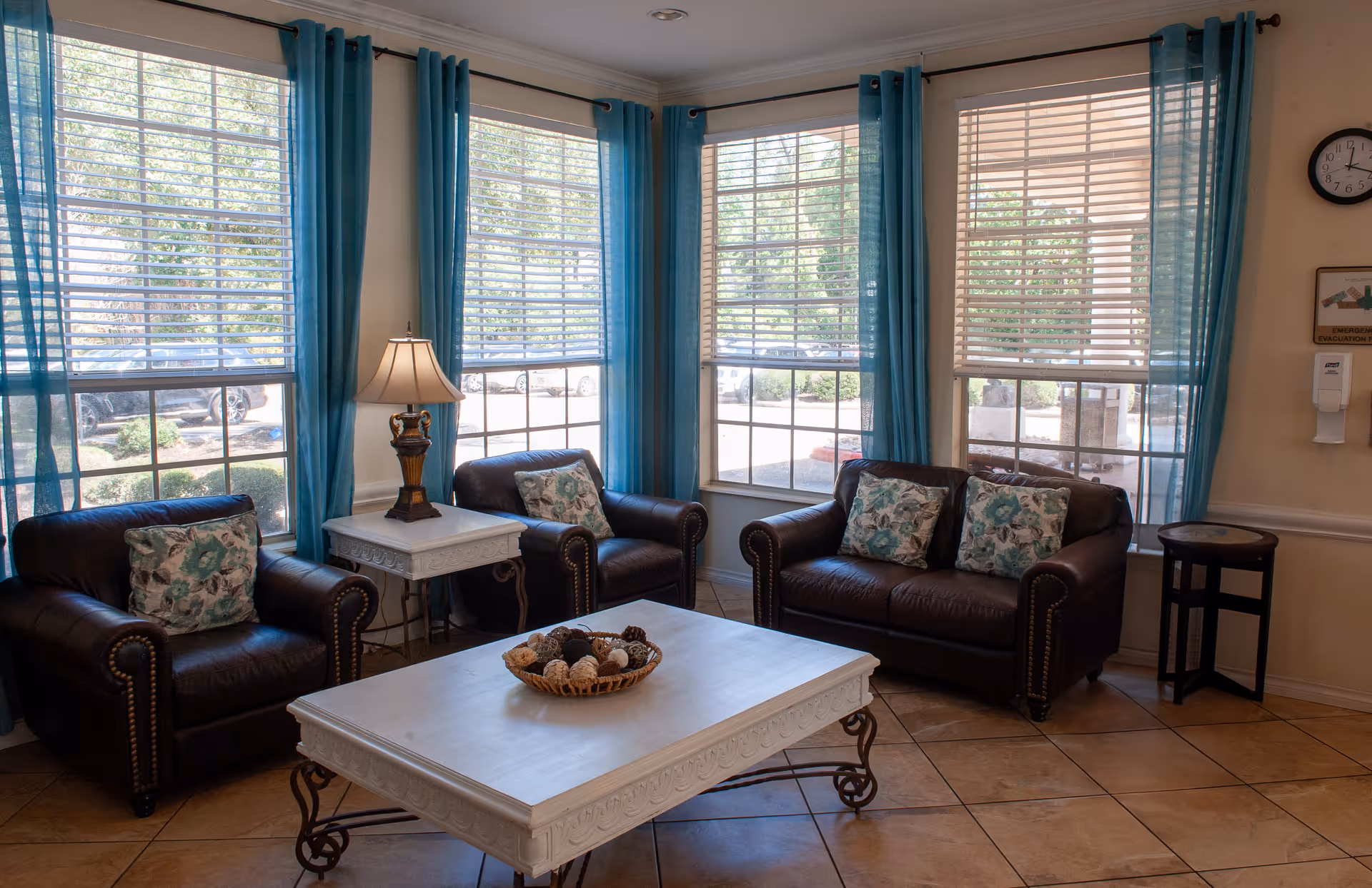 A cozy sitting area with three dark brown leather armchairs, each adorned with floral patterned cushions. The chairs are arranged around a white wooden coffee table with decorative legs, which holds a basket of assorted decorative balls. The room has large windows with white blinds and turquoise curtains, allowing natural light to fill the space. A table lamp sits on a small white side table between two of the chairs. The floor is tiled in a warm beige color.
