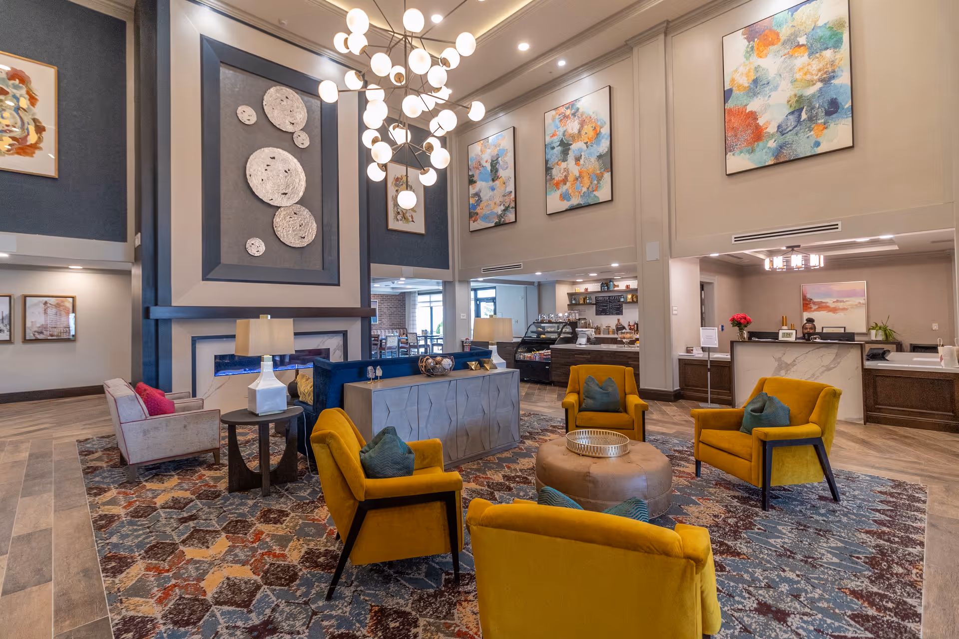 A spacious and stylish senior living facility common area with high ceilings, modern chandelier, and colorful abstract artwork on the walls. The seating area features mustard yellow armchairs with blue pillows arranged around a round ottoman on a patterned rug. There is a fireplace with decorative wall art above it, a reception desk in the background, and a small café or snack bar area with shelves and a display case.