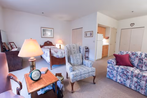 A cozy senior living room and bedroom area combined in one space. The room features a floral-patterned sofa with a red pillow, a blue and white patterned armchair, a wooden side table with a lamp and clock, and a bed with a floral bedspread against the back wall. There is a small kitchen area visible in the background with a refrigerator and cabinets. The walls are white, and the room has a carpeted floor.