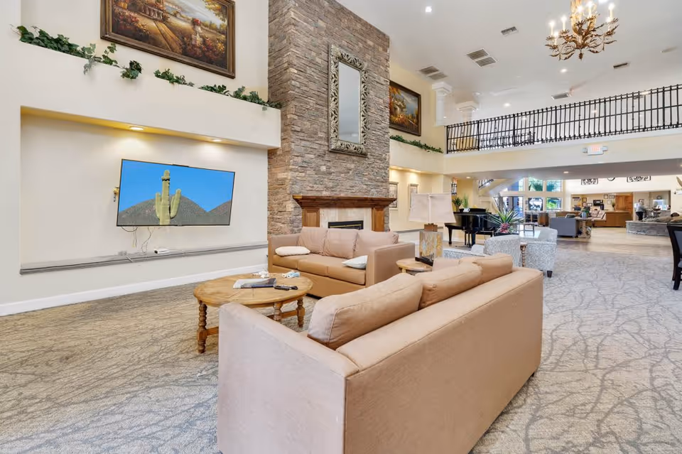 Spacious senior living facility common area with beige sofas, a round wooden coffee table, a stone fireplace with a decorative mirror above it, a wall-mounted TV displaying a cactus image, and a chandelier hanging from the ceiling. The room has high ceilings, carpeted floors, framed paintings, and a balcony railing overlooking the space.