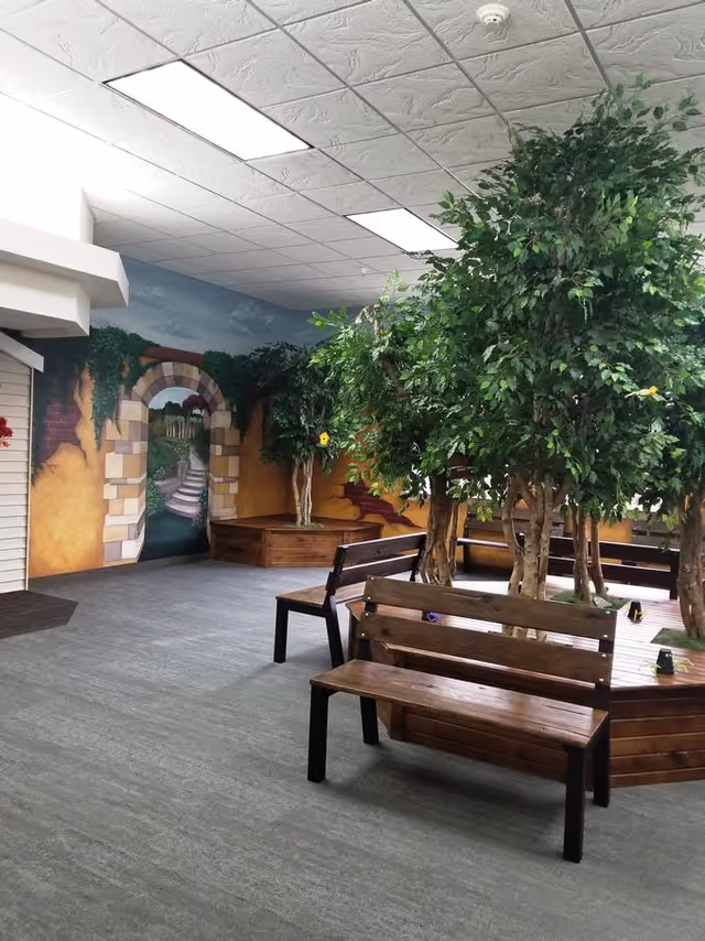 Indoor seating area with wooden benches surrounding planters that hold large leafy artificial trees. The walls are decorated with a mural depicting an archway leading to a garden path. The ceiling has white tiles with fluorescent lights.