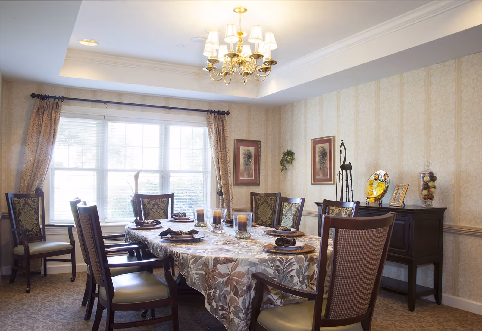 A well-lit dining room with a large rectangular table covered with a floral tablecloth. The table is set with plates, napkins, and candles. Surrounding the table are eight wooden chairs with cushioned seats and backs. The room has beige patterned wallpaper, a window with curtains, a chandelier hanging from the ceiling, and a dark wooden sideboard decorated with framed pictures, a sculpture, and a glass jar filled with decorative items.