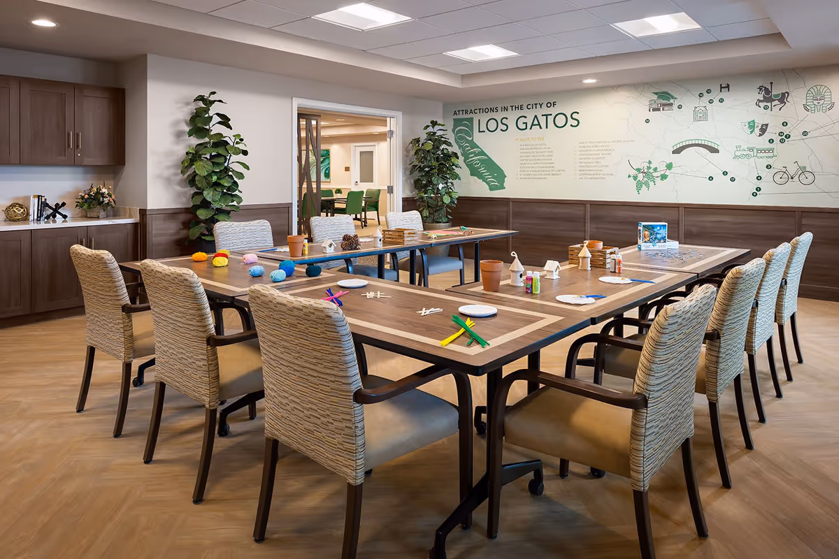 A well-lit activity room with a large U-shaped table surrounded by cushioned chairs. The table has various craft supplies including yarn, paint bottles, small wooden birdhouses, and pipe cleaners. The room features wood flooring, wooden cabinets on one side, and two large potted plants. On the wall, there is a map and text highlighting attractions in the city of Los Gatos, California. Through an open doorway, another room with green chairs and artwork is visible.