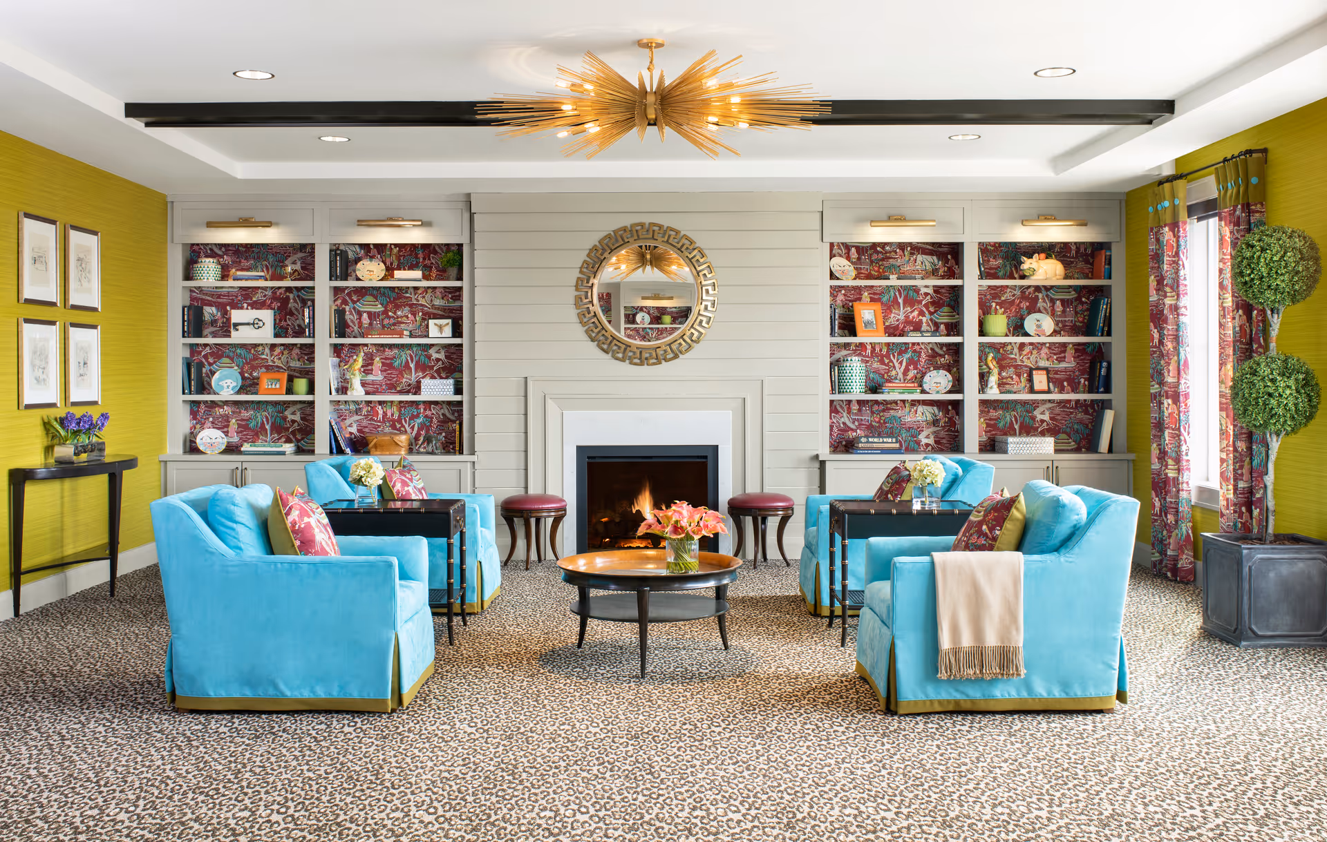 A bright and colorful living room with four turquoise armchairs arranged around a round wooden coffee table with a vase of flowers. The room features a fireplace with a round decorative mirror above it, built-in shelves with decorative items and books, yellow-green walls, patterned curtains, and a leopard print carpet. A modern gold chandelier hangs from the ceiling.