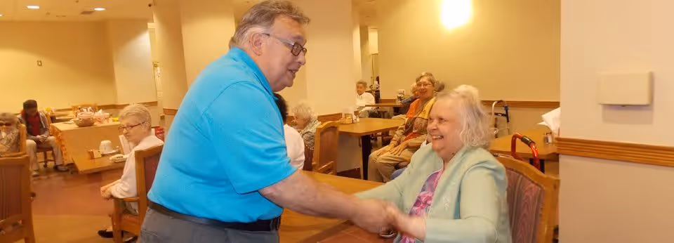 A man in a blue shirt is warmly shaking hands with an elderly woman seated at a table in a communal dining area. Several other elderly individuals are seated at tables in the background, engaging in conversation or activities. The room is well-lit with beige walls and wooden furniture.