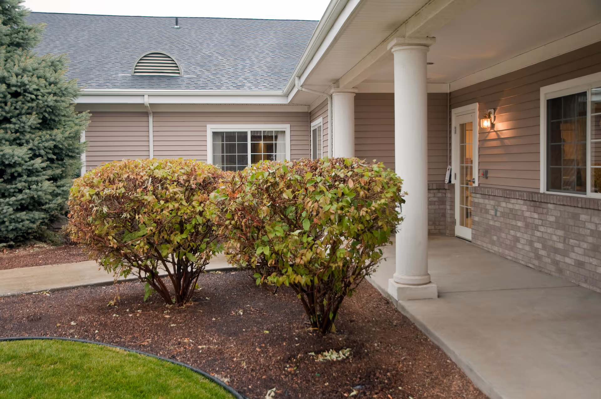 Exterior view of a building with beige siding and brick accents, featuring a covered walkway supported by white columns. There are trimmed bushes and a tree in the landscaped area next to the walkway, and a lit wall-mounted light near a glass door.