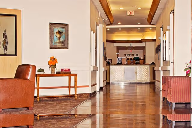 A well-lit interior hallway of a senior living facility with polished wooden floors, a reception desk at the far end, and seating areas with brown and red chairs on either side. There is a wooden table with a flower vase and a book on it, and framed artwork on the walls.