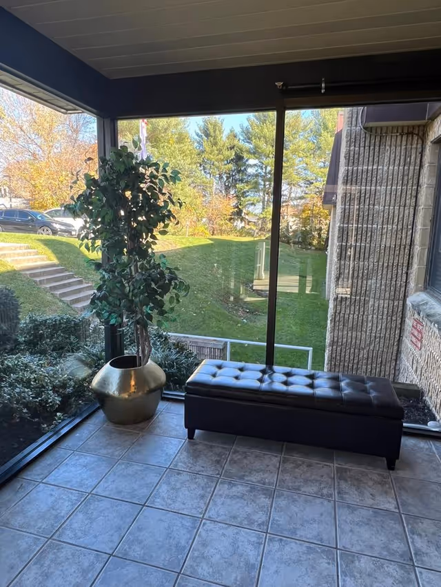 A small indoor seating area with a black tufted bench and a large potted plant in a gold-colored pot. The area has tiled flooring and large glass windows providing a view of green grass, bushes, trees, and parked cars outside.