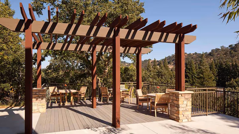 Outdoor patio area with wooden pergola, several tables and chairs, surrounded by trees and hills in the background under a clear blue sky.