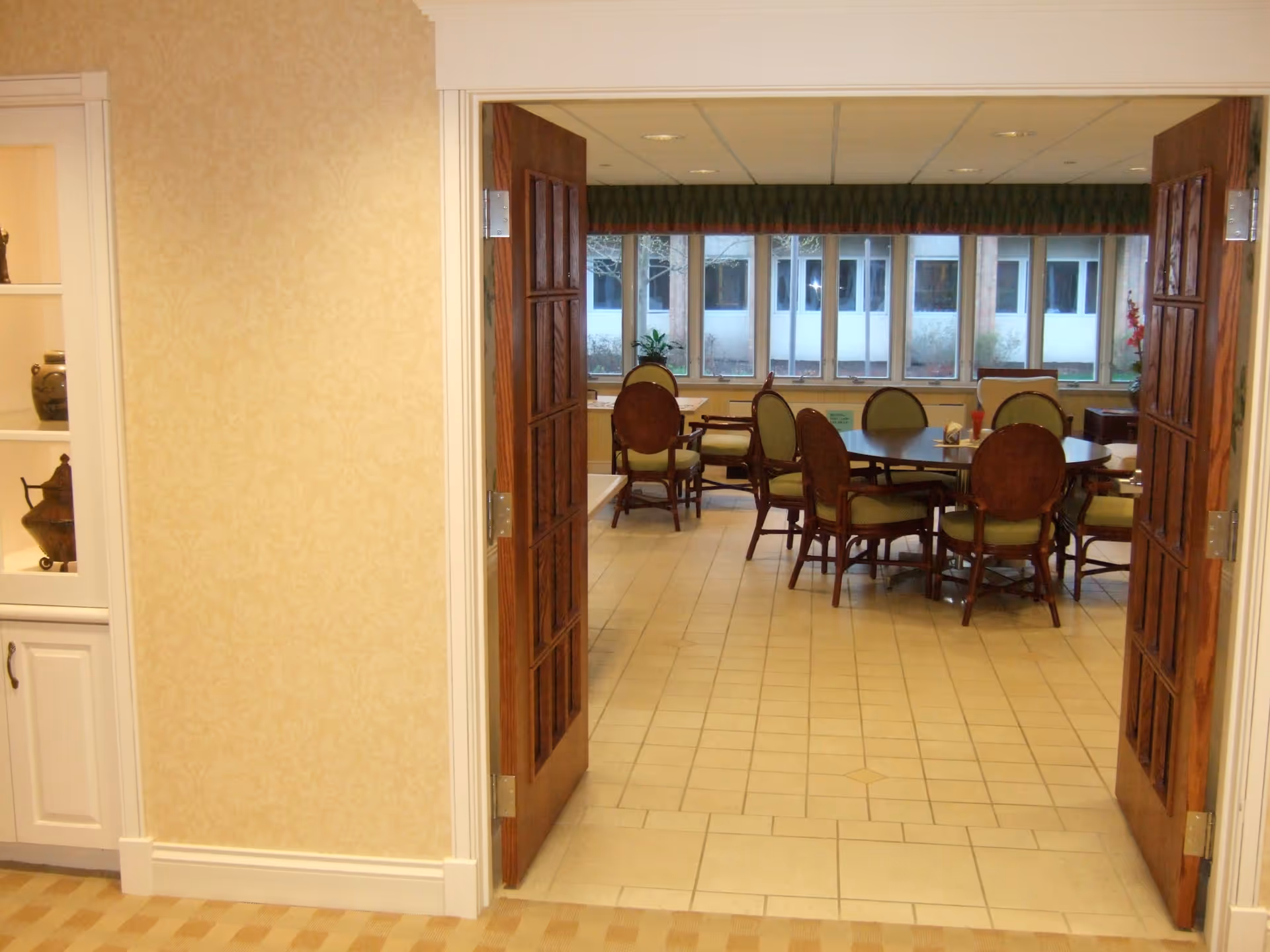 Open wooden double doors leading into a dining area with round tables and green-upholstered chairs and windows along the back wall.