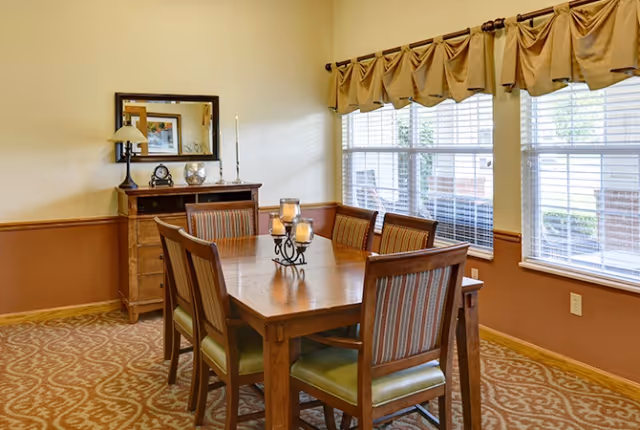 Dining room with a wooden table and six chairs, a candle centerpiece, sideboard, and large windows with valances.