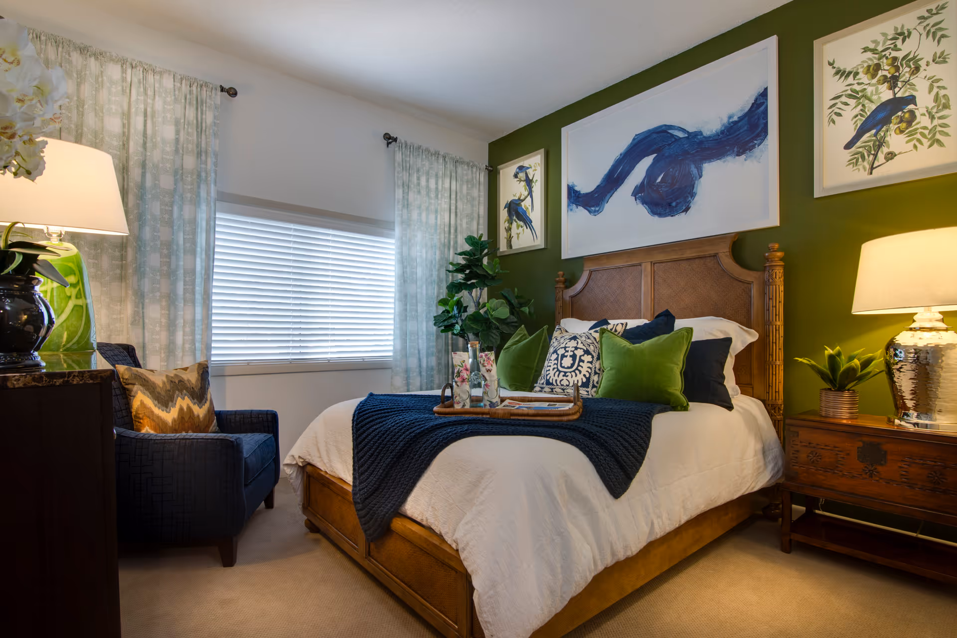 A cozy bedroom with a wooden bed frame, white bedding, and a dark blue throw blanket. The bed is decorated with green and blue pillows. There is a wooden nightstand with a lamp and a small plant on the right side of the bed. On the left side, there is a dark blue armchair with a patterned pillow. The walls are painted green behind the bed and white elsewhere, adorned with framed artwork featuring birds and abstract designs. Light blue patterned curtains frame a window with closed blinds.