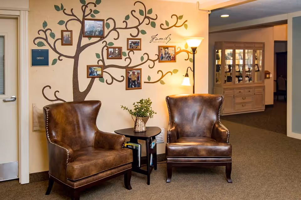 Two brown leather armchairs with nailhead trim are positioned around a small black side table holding a vase with greenery. Behind the chairs, a wall features a painted tree with framed family photos hanging on its branches and the word 'Family' written on the wall. To the left, there is a door labeled 'Office,' and to the right, a wooden cabinet with glass doors is visible in a hallway.