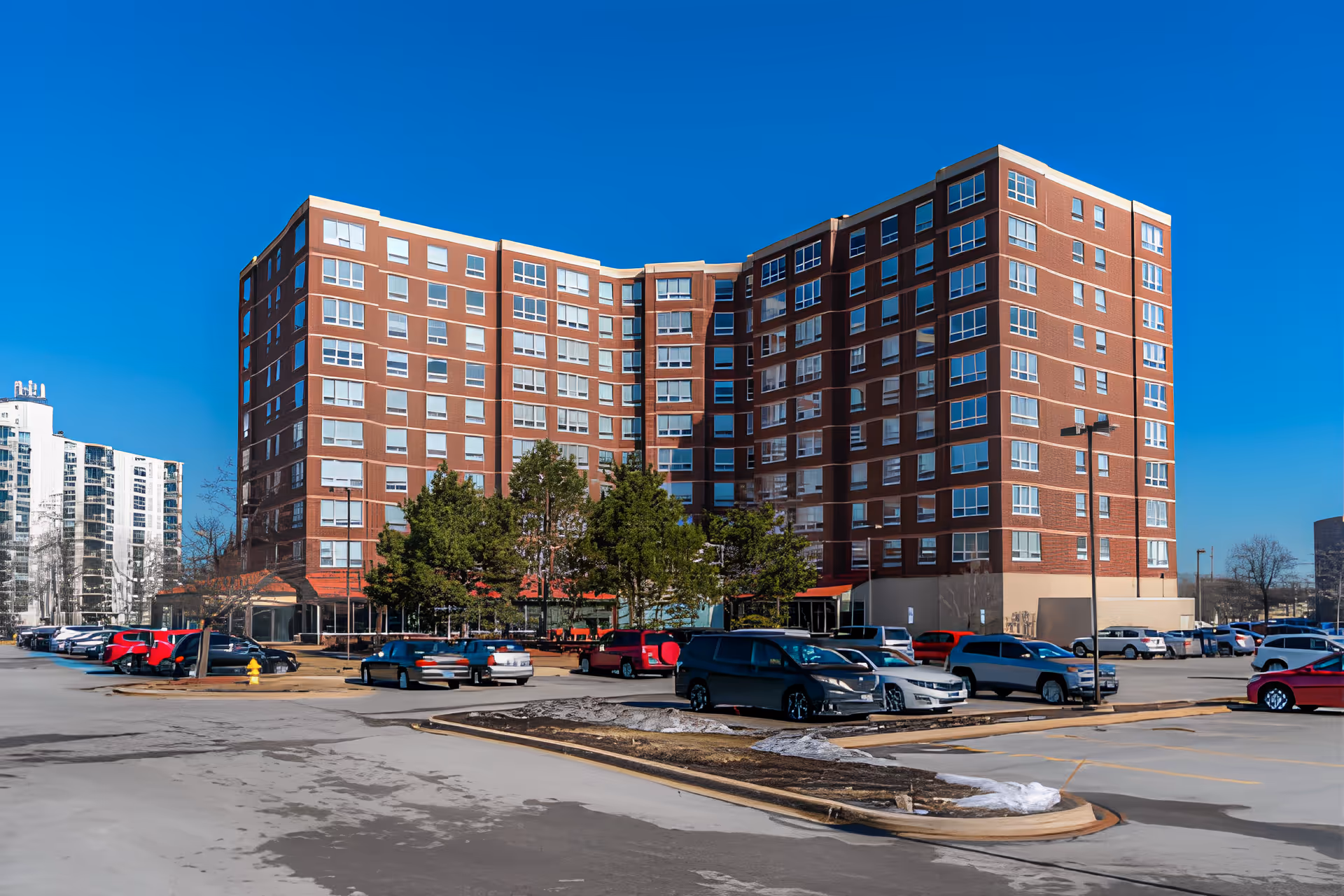 Exterior view of a large multi-story brick building with many windows, surrounded by a parking lot with several parked cars and some trees in front. The sky is clear and blue.