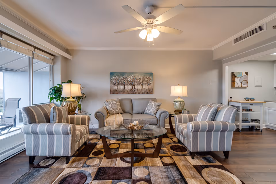 A cozy living room with a gray sofa and two striped armchairs arranged around a glass coffee table on a patterned rug. Two table lamps with beige shades sit on side tables next to the sofa. A ceiling fan with lights is mounted on the ceiling. A large window with blinds lets in natural light, and a painting of trees hangs on the wall behind the sofa. The room has wooden flooring and an open doorway leading to a kitchen area.