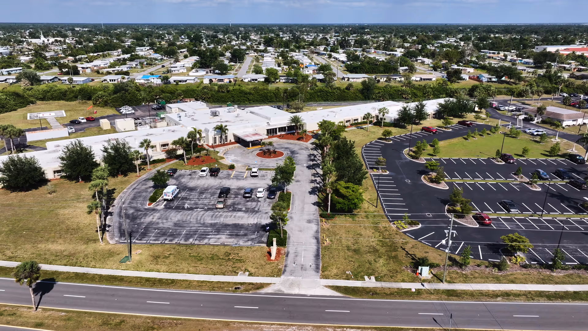 Aerial view of Village Place Health and Rehabilitation Center showing a single-story building with two parking lots, surrounded by grass and trees, with a residential neighborhood in the background.
