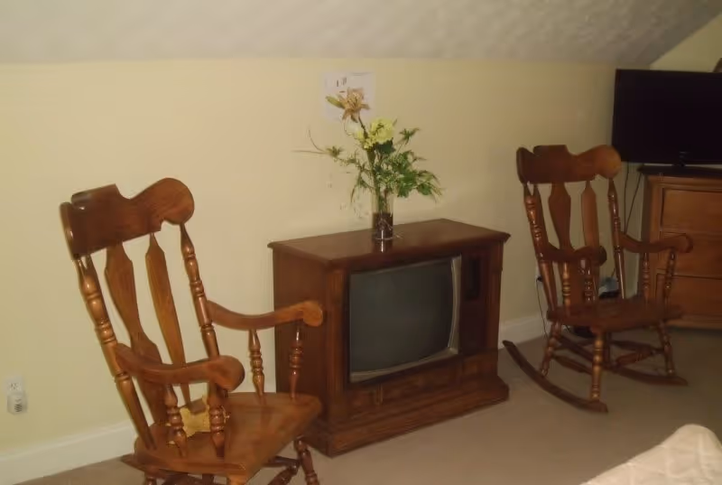 A small room with two wooden rocking chairs facing each other, a vintage wooden TV cabinet with an old television set on top, and a vase with flowers placed on the cabinet. A modern flat-screen TV is visible on a wooden dresser in the background.