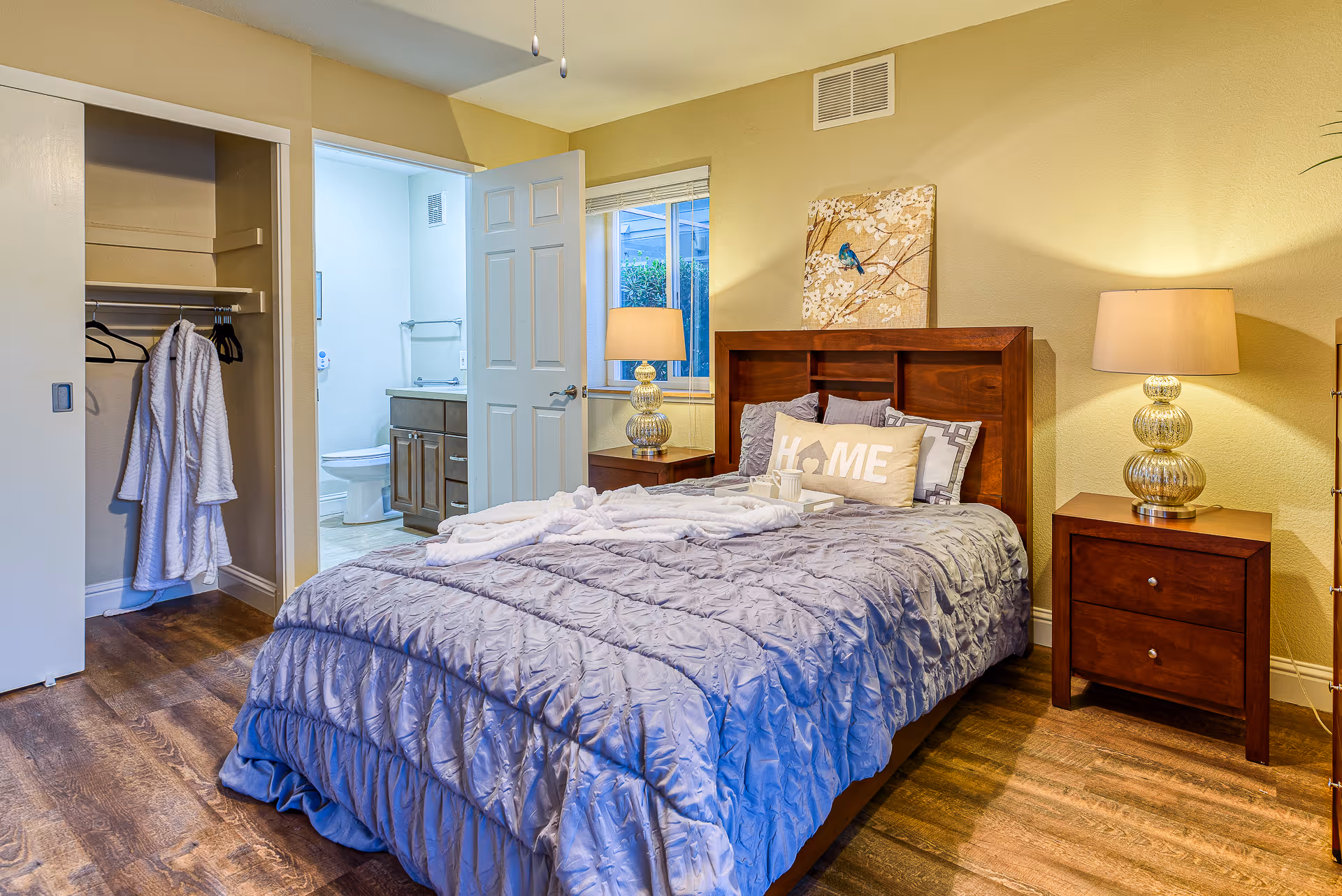 A furnished bedroom with a wooden bed and nightstands, an open closet with robes, and a doorway leading to an en-suite bathroom.