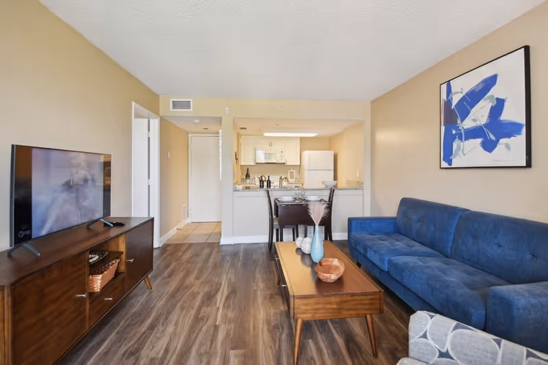 A modern living room with a blue sofa, wooden coffee table with decorative items, and a TV on a wooden stand. The room has wood flooring and beige walls with a blue abstract painting. In the background, there is a small dining area with a table and chairs, and a kitchen with white cabinets, a refrigerator, and a microwave.