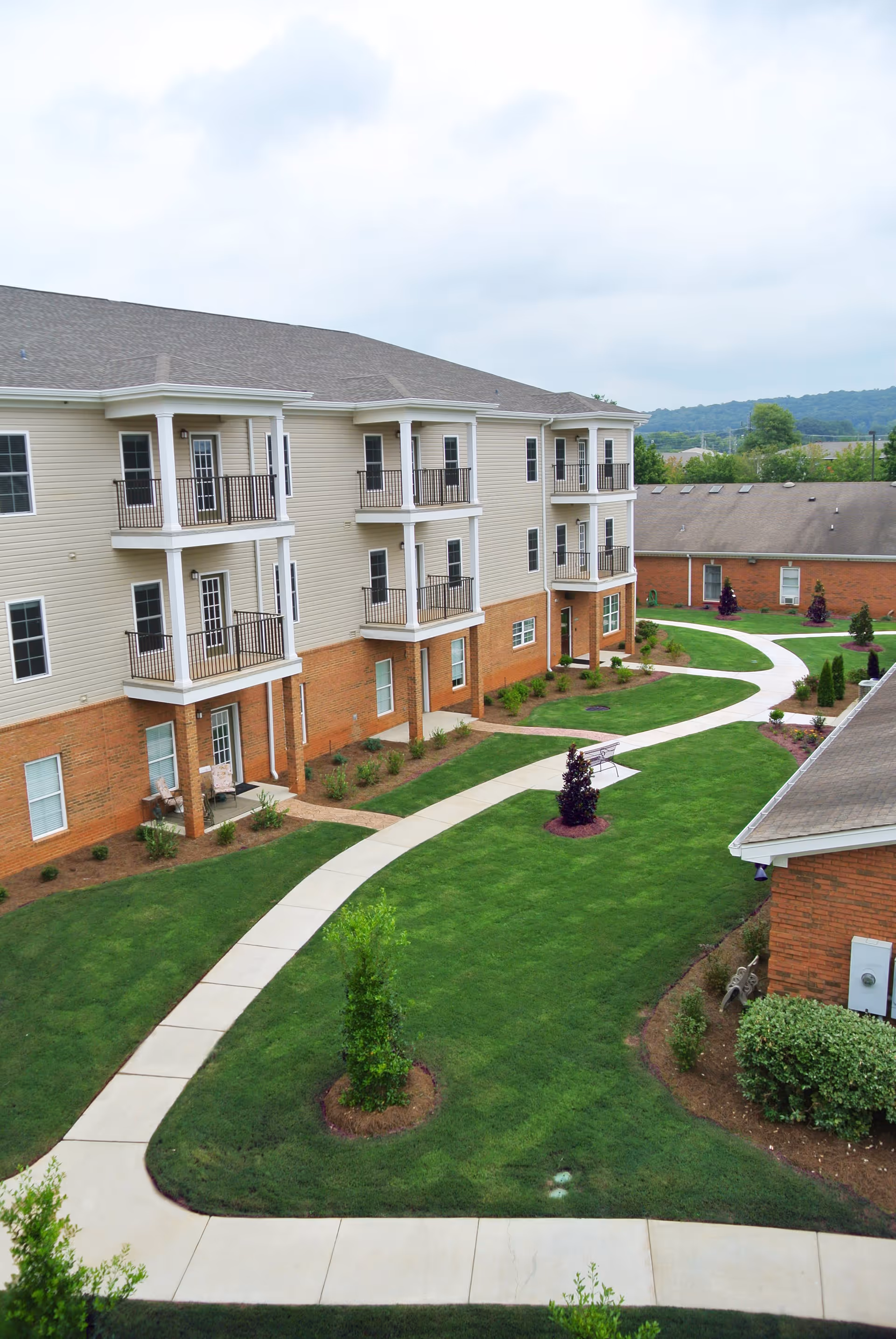 View of a three-story residential building with balconies overlooking a well-maintained green lawn and winding concrete pathways in an outdoor courtyard area.