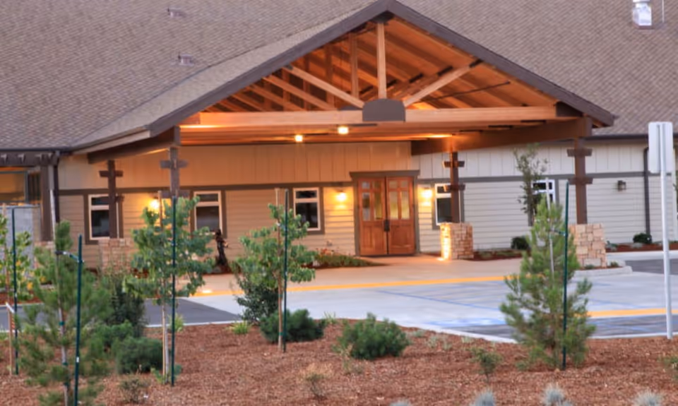 Entrance of a single-story memory care residence with a covered wooden porte-cochère, double doors, exterior lights, and landscaped front area.