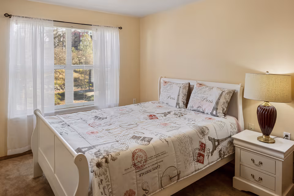 A cozy bedroom with a white wooden bed frame and a bed covered with a Paris-themed quilt and matching pillows. There is a white nightstand with two drawers next to the bed, topped with a red lamp with a beige lampshade. A large window with sheer white curtains lets in natural light, showing trees outside.