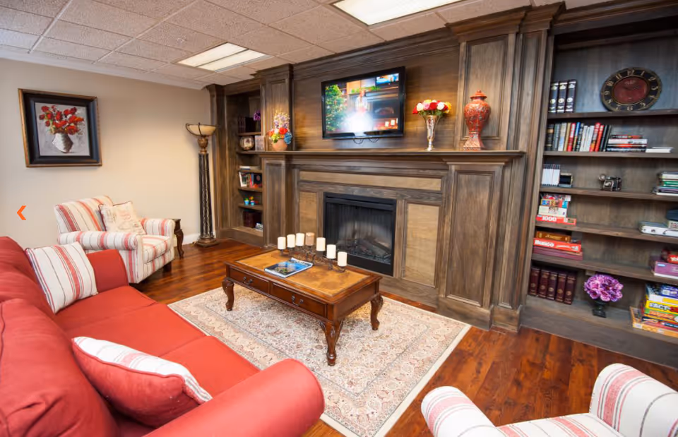 Cozy living room with a red sofa and striped armchairs surrounding a wooden coffee table on a patterned rug. The room features a large wooden built-in bookshelf and fireplace with a mounted TV above it. Decorative items and books are arranged on the shelves, and a painting hangs on the wall.
