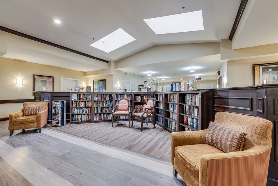 A cozy reading area in a senior living facility featuring bookshelves filled with books, two upholstered armchairs with patterned cushions, and two wooden chairs with cushions. The space is well-lit with ceiling lights and natural light from skylights above. The walls are painted in a soft beige color with framed artwork, and the floor has a combination of wood and carpet.