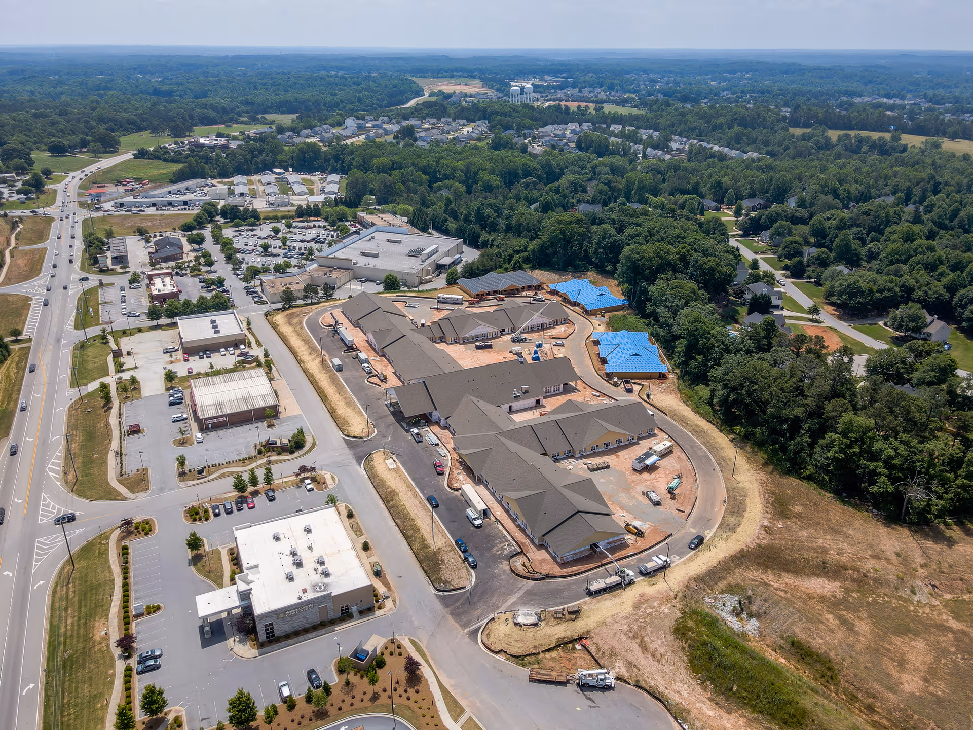 Aerial view of Manor Lake Assisted Living & Memory Care - Hoschton under construction, surrounded by roads, parking lots, and greenery with residential neighborhoods in the distance.