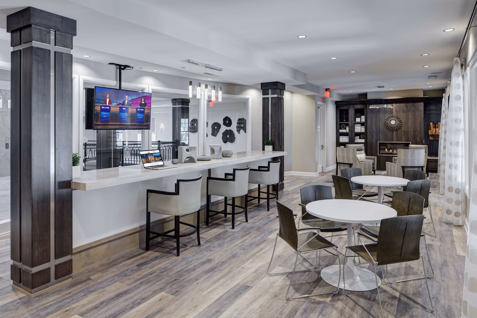 A modern common area with a long white counter and several cushioned chairs on one side. There are two round tables with dark chairs in the foreground. The room features wood flooring, dark wooden pillars, and a wall-mounted TV. In the background, there is a cozy seating area with armchairs and a fireplace, along with built-in shelves and decorative items.