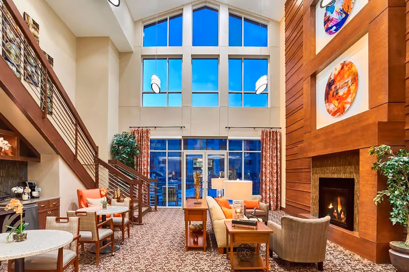 Spacious two-story senior living lounge with tall windows, multiple seating areas, small dining tables, and a fireplace.