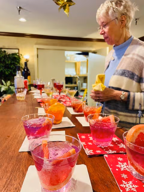 An elderly woman standing at a wooden counter with multiple colorful drinks in clear glasses arranged on napkins. The setting appears to be indoors with warm lighting and a cozy atmosphere.