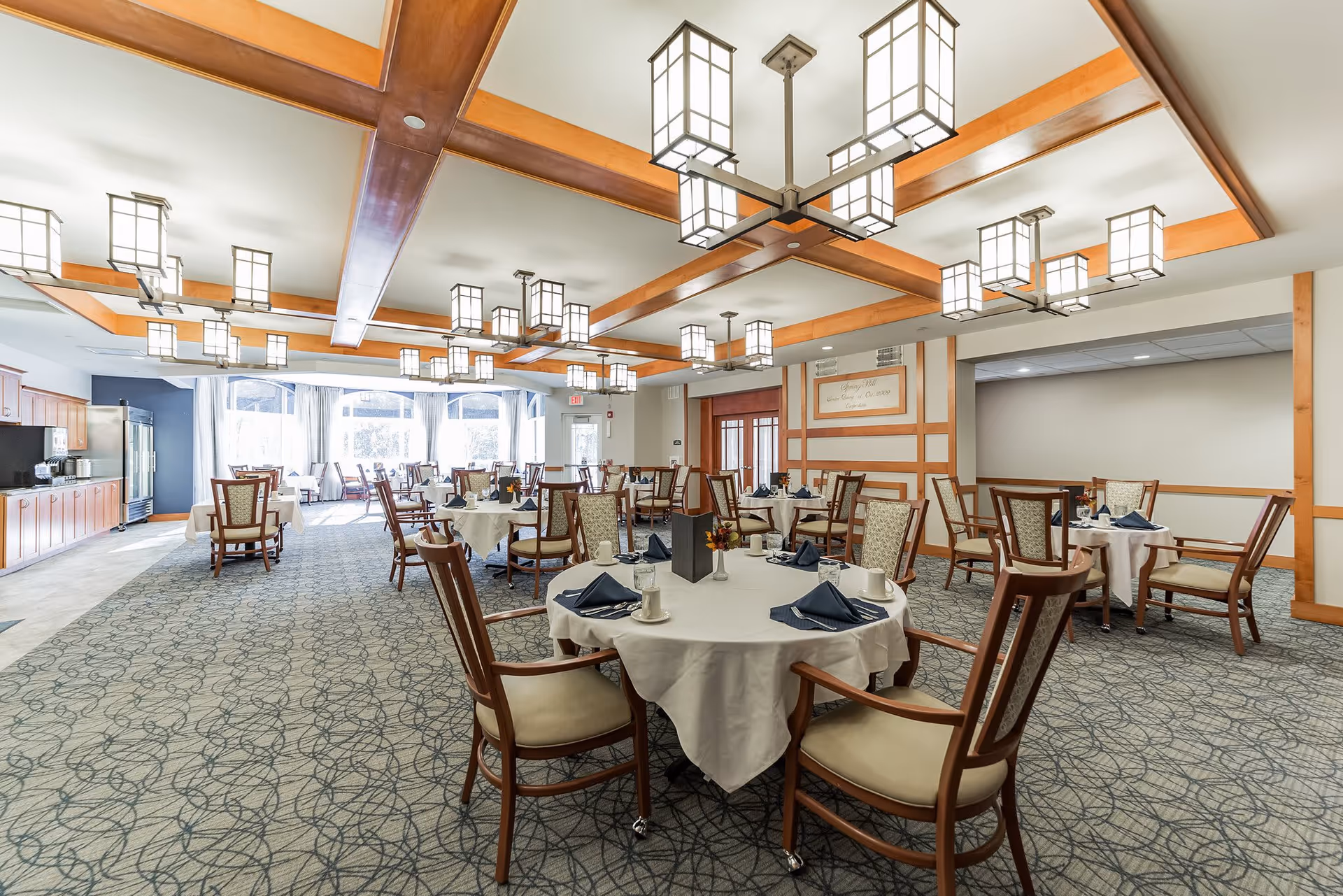 A spacious dining room with round tables covered in white tablecloths, each set with blue napkins, cups, and small flower arrangements. The room features wooden beams on the ceiling with multiple modern square pendant lights. Large windows at the far end allow natural light to fill the space. Chairs with cushioned seats and backs surround the tables, and there is a counter with cabinets and a refrigerator along one wall.