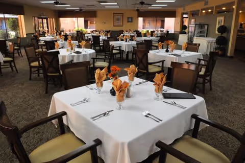 Spacious dining room with multiple tables covered in white tablecloths set with folded orange napkins and chairs.