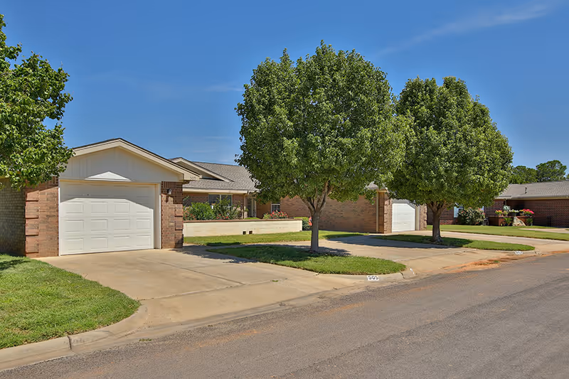 Exterior view of a single-story brick residential building with a white garage door, two green leafy trees in front, and a clear blue sky above.