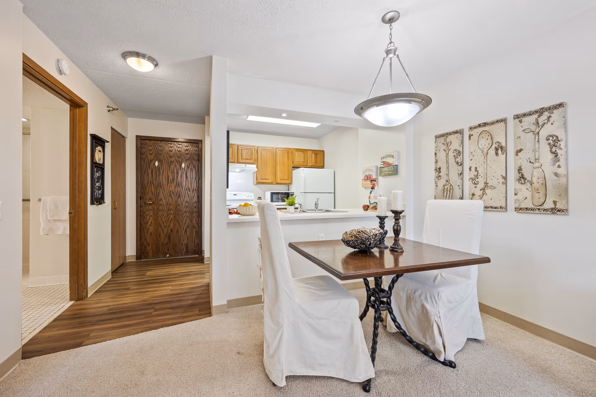 A dining area in a senior living facility featuring a wooden table with two white slipcovered chairs. The table is decorated with a candle holder and a decorative bowl. Behind the dining area is a kitchen with wooden cabinets, a white refrigerator, stove, and microwave. The walls are adorned with artwork depicting vintage kitchen utensils. The floor transitions from carpet in the dining area to wood in the hallway leading to a closed wooden door and a bathroom with a tiled floor.