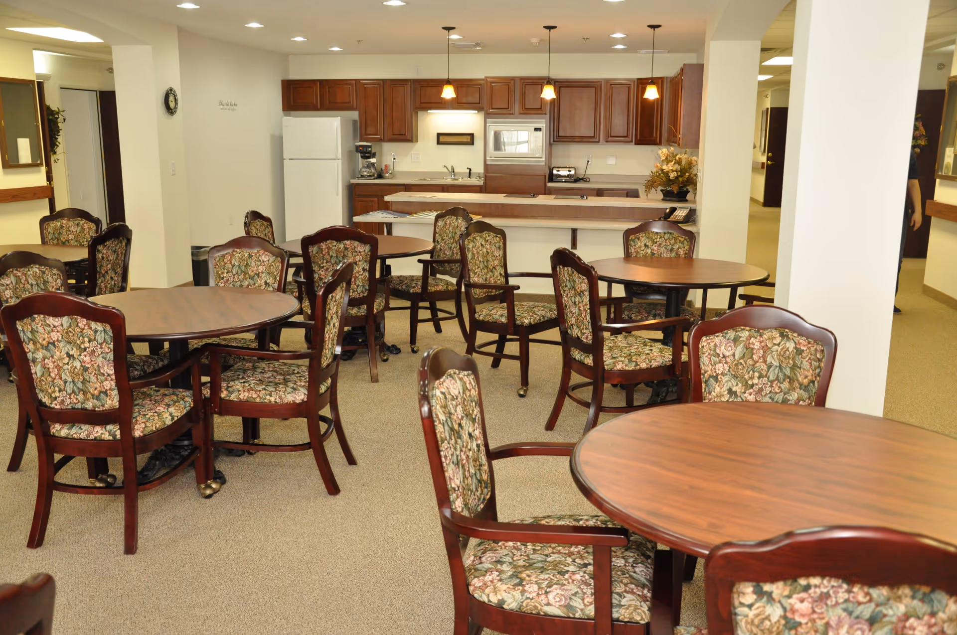 A dining area in an assisted living community featuring several round wooden tables surrounded by floral upholstered chairs. In the background, there is a kitchen area with wooden cabinets, a white refrigerator, a microwave, and a coffee maker. The room is well-lit with ceiling lights and pendant lights above the kitchen counter.