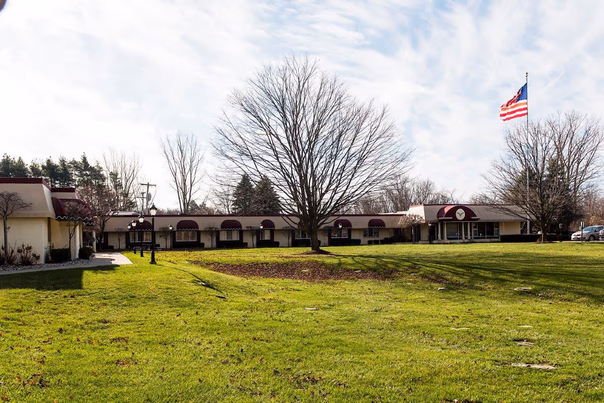Exterior view of a single-story building with maroon awnings over the windows and entrances, surrounded by a large grassy lawn with a few leafless trees. An American flag flies on a tall flagpole to the right side of the building under a partly cloudy sky.